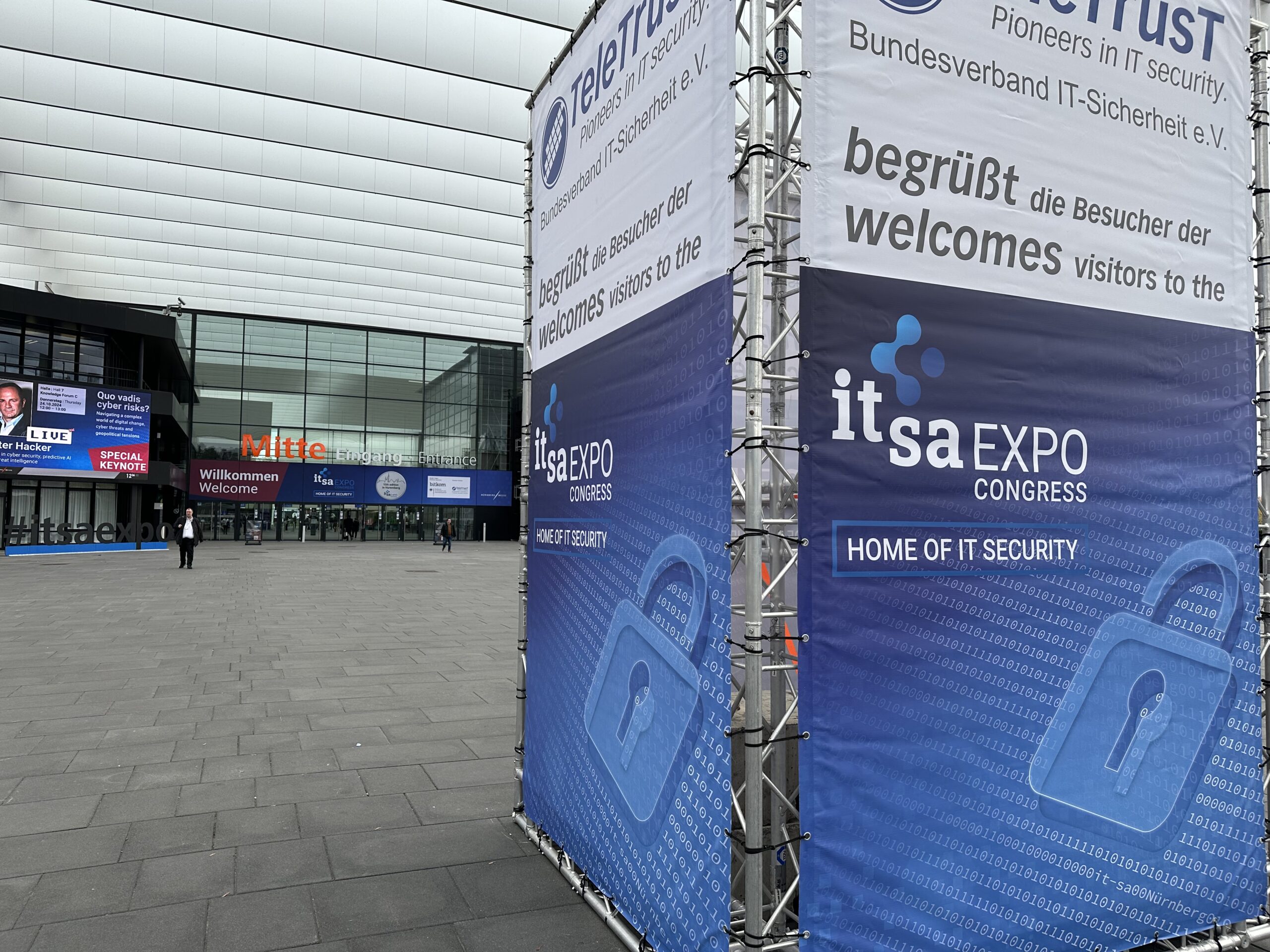 Eingang zum itsa Expo Congress mit Schildern mit einem blauen Vorhängeschloss-Logo. Die Banner heißen die Besucher der Veranstaltung willkommen und betonen den Schwerpunkt auf IT-Sicherheit. Die Expo findet in einem modernen Gebäude mit großen Glastüren statt.